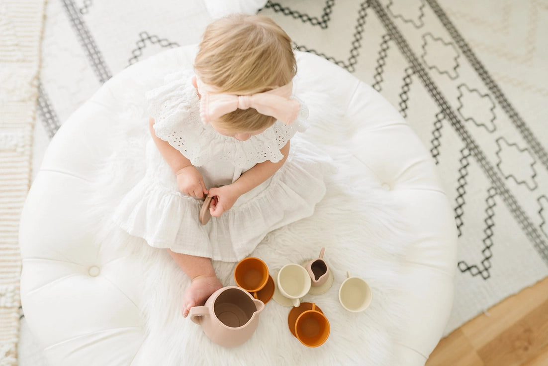 Little toddler girl wearing a white dress and playing with the silicone tea cup play set on a white carpet
