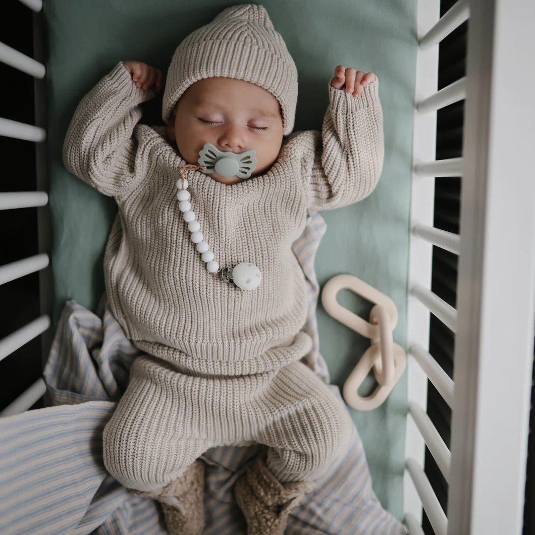 Baby sleeping in his crib with a pacifier made of silicon and wearing a chunky knitwear ensemble for babies in a beautiful color grey